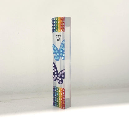 May include: A clear acrylic mezuzah case with rainbow-colored beads at the top and bottom. Two butterfly designs are painted on the front, one light blue and one dark blue. A Hebrew letter shin is visible above the light blue butterfly.